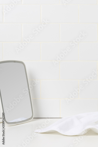 Minimalist beauty and skincare routine mockup background. White tabletop mirror and soft cotton towel on a counter against white subway tile wall with copy space.
