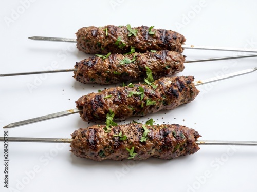 Four freshly grilled Turkish kofta kebabs on metal skewers garnished with green herbs, isolated on a bright white background, ideal for Middle Eastern restaurant menu design and food blogs.