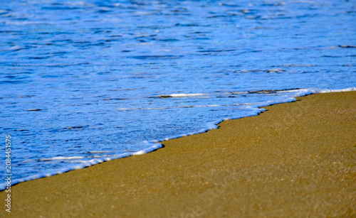 Fotografie A delicate wash of shallow blue sea water creating a thin elegant line of white foam across the smooth sandy beach defining the exact boundary of the peaceful coastal shoreline