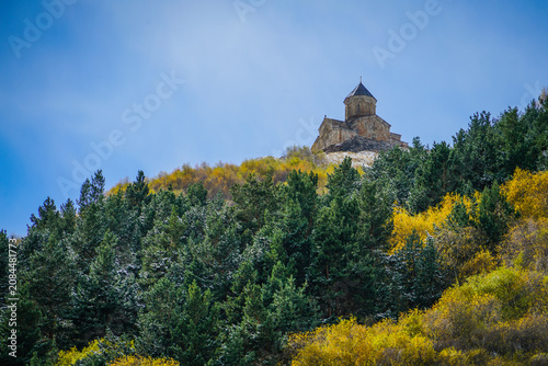 Gergeti Trinity Church, Stepantsminda, Georgia, overlooking a vibrant autumn forest beneath a clear blue sky.