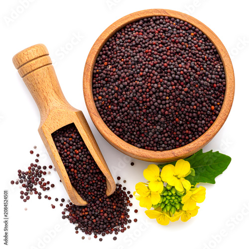 Top view of wooden bowl and scoop of black mustard seed with green leaf on white background