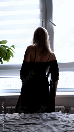 A sexy beautiful woman in a black erotic negligee stands near the window in the bedroom.
