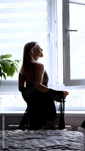 A sexy beautiful woman in a black erotic negligee stands near the window in the bedroom.