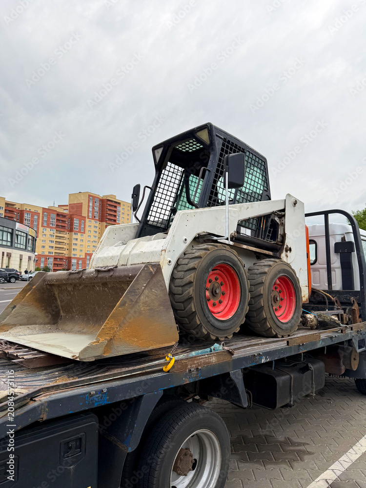 Obraz premium Compact skid steer loader with a bucket attachment parked on a flatbed truck in an urban environment with buildings visible in the background