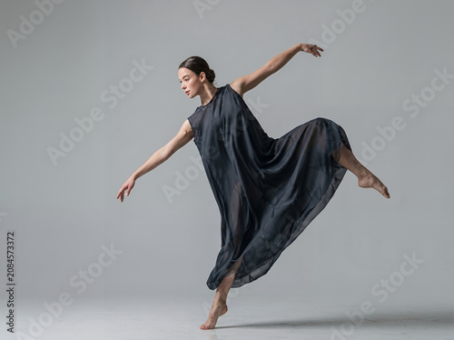 Graceful professional ballerina dancing in a flowing elegant dark grey dress, performing expressive contemporary ballet dance in studio isolated on grey backdrop. Concept of fine art and movement.
