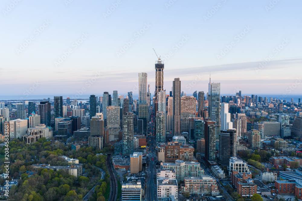 Obraz premium Aerial skyline view over downtown Toronto showing dense modern high rises, central tower under construction, waterfront horizon, parks, streets, and warm evening light