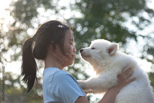 A happy Asian girl looking affectionately at a cute fluffy white puppy, outdoor natural light, friendship and love concept.