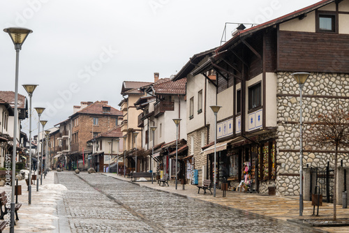 A quiet, wet cobblestone street lined with traditional stone and wood buildings in the historic old town, on a cloudy winter day in Bansko, Bulgaria.