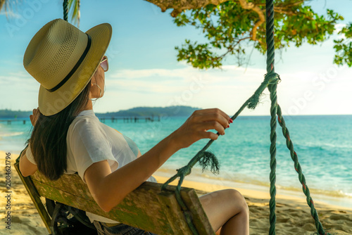 Tableau sur toile Woman relaxing on a wooden beach swing beneath a tropical tree while overlooking turquoise ocean