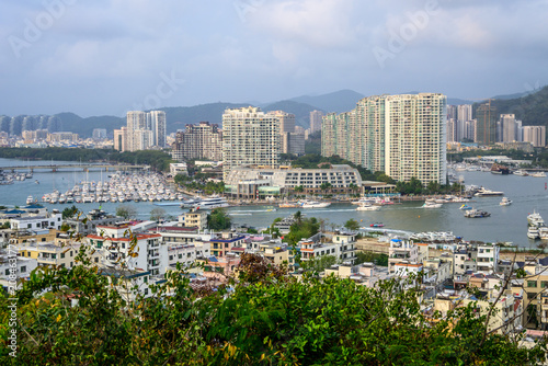A panoramic view of a Sanya, Hainan, China with high rise buildings, a bustling marina filled with boats, and a lush green foreground. The cityscape extends towards mountains in the background.