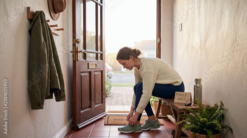 A woman sits on a wooden bench in a hallway, tying her green sneakers next to a card that reads 
