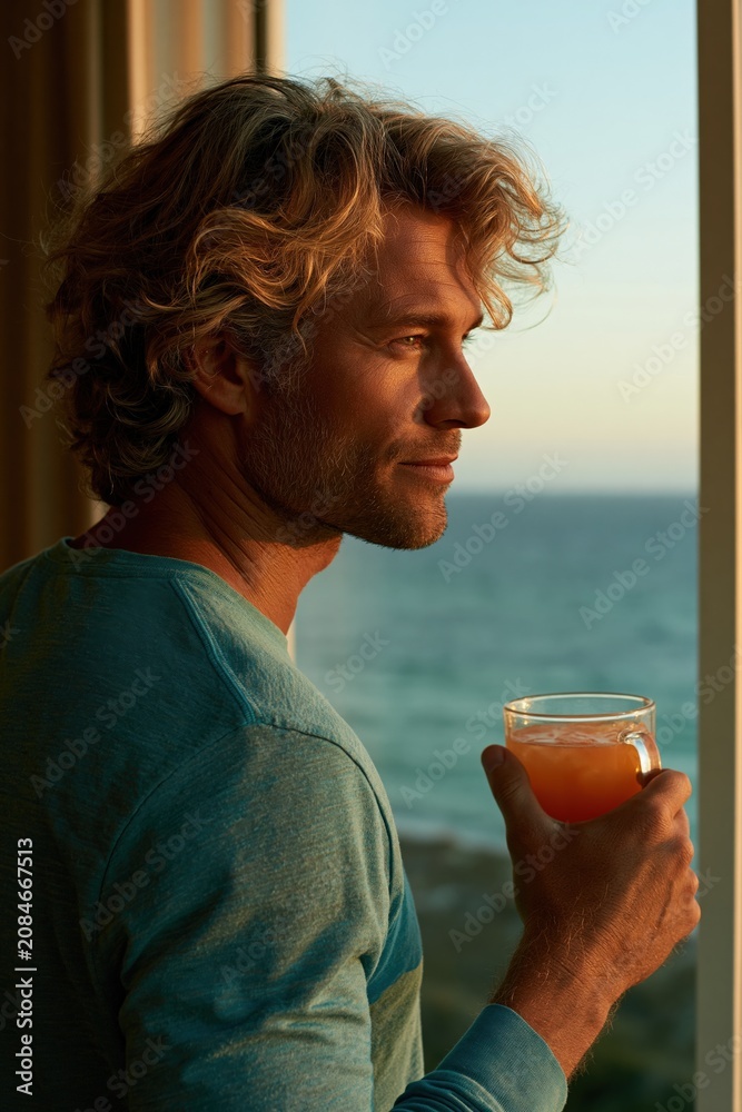 Fototapeta premium Side profile man with a glass of juice looking out to the ocean at sunset for seaside lifestyle background and contemplative banner