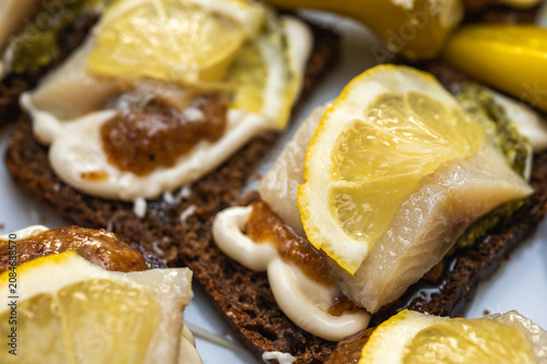Close-up of gourmet seafood appetizers with salted fish, lemon slices, and green pesto on dark rye bread. Traditional European snacks on white plate. Macro view, selective focus, blur