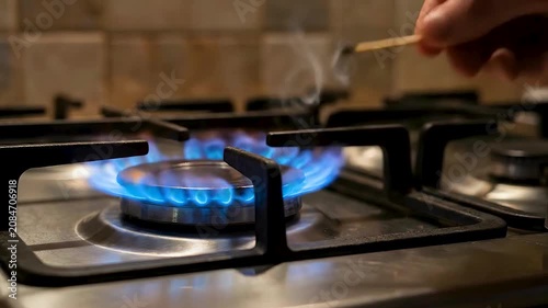 A hand is holding a lit match to a blue gas flame on a stovetop. The match is igniting the gas, creating orange flames.