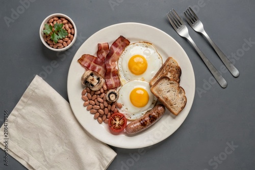 Full English Breakfast with fried eggs, bacon, sausages, kidney beans, bread, linen napkin, concept of hearty meal featuring eggs, bacon, sausage, beans, mushrooms, toast.
