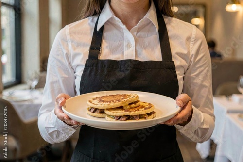 Woman in white shirt, black apron, holding lokse filled with duck fat, smiling. Concept of cozy restaurant atmosphere featuring delicious potato flatbread with duck fat filling.