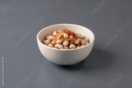 Prebranac in small white bowl, filled with round white beans topped with orange onion. Concept of baked beans with onions paprika oil, highlighting flavors and textures.