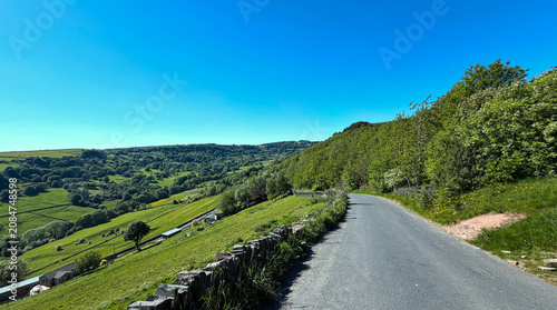 Howes Lane meanders through lush green hills under a clear blue sky. The serene landscape offers a peaceful countryside view, perfect for a leisurely drive in Shibden, Halifax, Yorkshire, UK