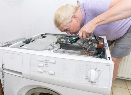 Fotografía Housewife uses a flashlight to carefully inspect the wiring inside a disassembled, faulty washing machine to diagnose the problem and call a repairman to the house