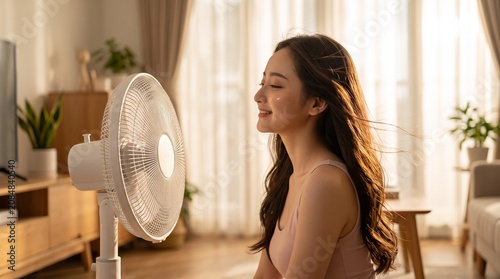 Happy Young Asian Woman Cooling Off in Front of Electric Floor Fan in Sunlit Living Room During Summer