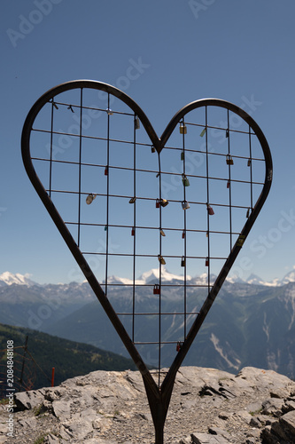 Photography Vertical shot of a large metal heart sculpture filled with love padlocks on top of a mountain cliff, overlooking scenic snow-capped alpine peaks and blue sky at Gemmi Pass, Switzerland