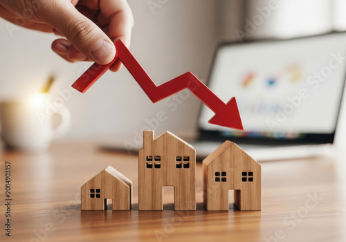 Red arrow pointing down over small wooden house models on office desk representing falling property values real estate crisis and recession.