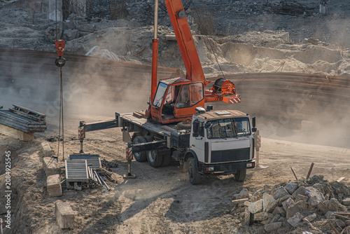 Construction scene featuring mobile crane mounted on truck, actively lifting heavy load, kicking up dust and debris. Truck with flatbed trailer equipped for heavy-duty work. Uneven ground with piles