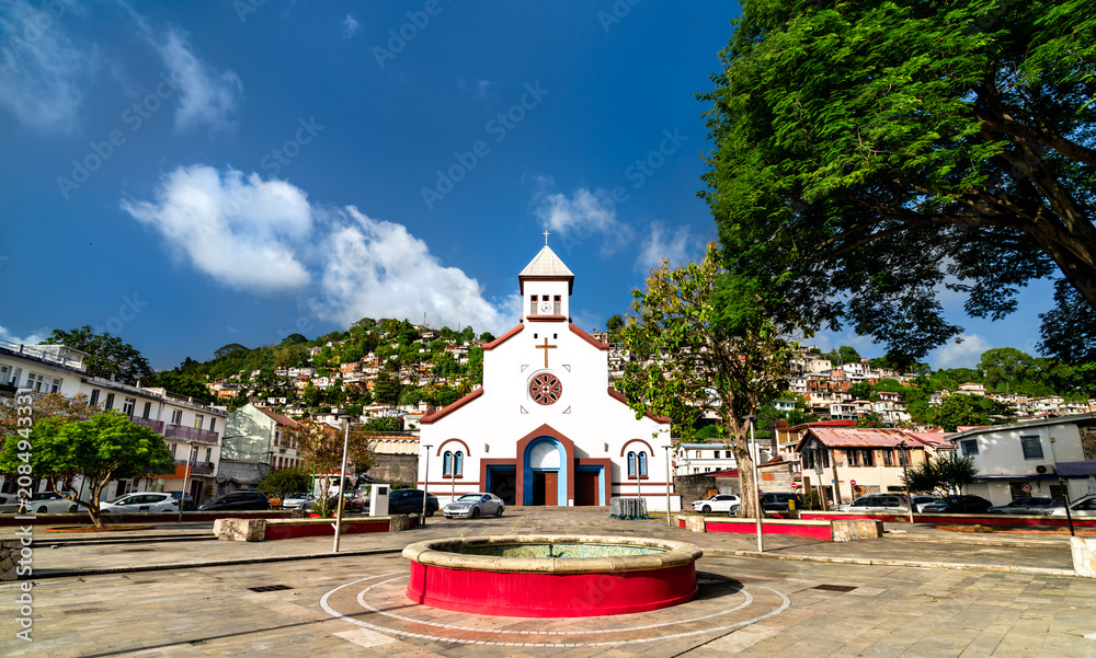 Fototapeta premium The white facade of Saint Anthony of Padua Catholic Church in Fort-de-France, Martinique with colorful hillside houses in the background under a blue sky.