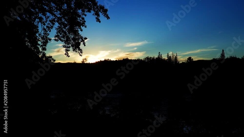 The last rays of a summer sunset illuminate a large Canadian river as the water flows gently through the darkening landscape. Warm golden colors contrast beautifully with the deep blue tones of approa