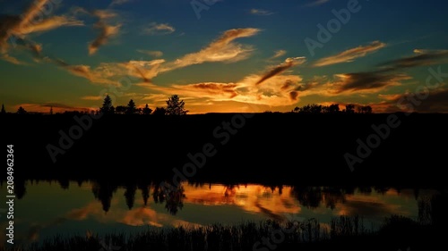 A breathtaking summer sunset reflects across the calm waters of a Canadian lake. Golden, orange, and warm evening colors create a peaceful landscape as the sun slowly disappears below the horizon.