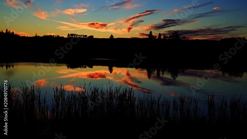 A breathtaking summer sunset reflects across the calm waters of a Canadian lake. Golden, orange, and warm evening colors create a peaceful landscape as the sun slowly disappears below the horizon.