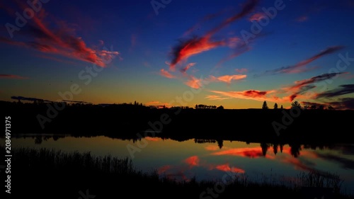 A breathtaking summer sunset reflects across the calm waters of a Canadian lake. Golden, orange, and warm evening colors create a peaceful landscape as the sun slowly disappears below the horizon.