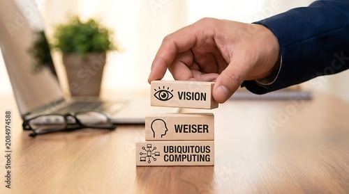Hand stacking wooden blocks with the words vision wiser and ubiquitous computing on a desk with a laptop
