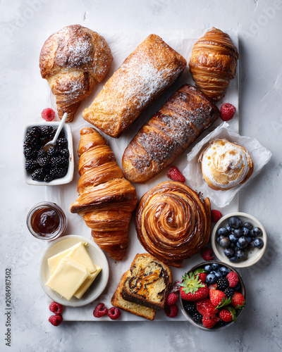 Top-down flat lay of artisan bakery assortment including croissants, sourdough bread, baguettes, cinnamon rolls, pain au chocolat, butter, jam and fresh berries on white stone background