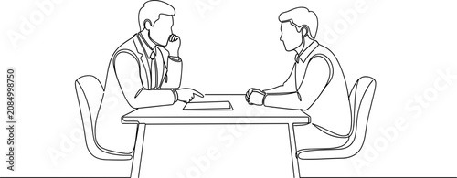 Two businessmen talking at a table, one pointing at a document.