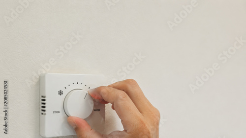 Hand turning white thermostat dial on wall, adjusting indoor temperature for home comfort, close up view of finger rotating round control knob