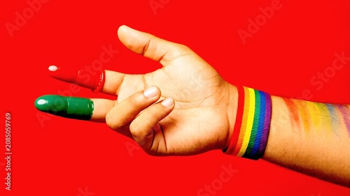 A hand wearing a rainbow pride wristband holds red and green condoms on its fingers against a vivid red background, promoting lgbtq+ safe sex, human rights, and diversity.