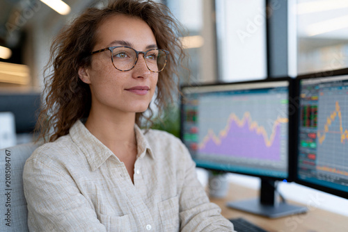 Young woman sits focused before dual monitors. Glasses rest gently on her nose, eyes calm, attentive. Charts pulse behind her-colors shift, trends move, data streams. She's immersed in analysis