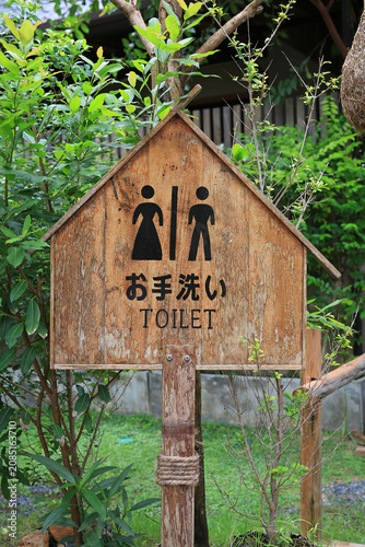 A rustic wooden sign with Japanese characters and clear symbols points the way to the restroom amidst a peaceful green garden.