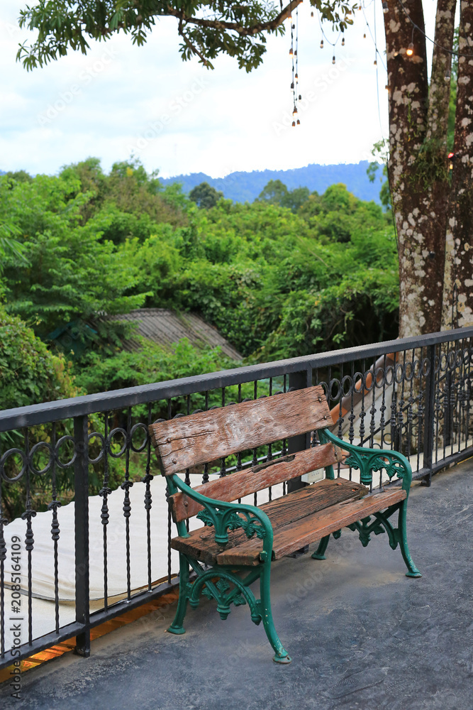 Fototapeta premium A weathered wooden bench sits quietly on a balcony, offering a peaceful spot to rest and enjoy the lush green forest view and the distant blue mountains.