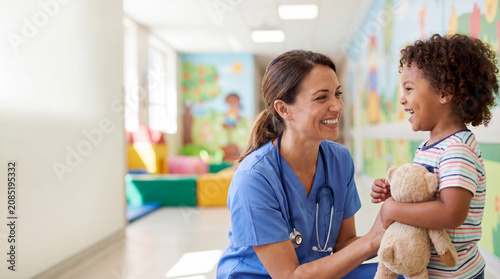 Smiling female healthcare professional interacting with a happy child holding a teddy bear in a colorful pediatric clinic hallway, professional medical care concept.