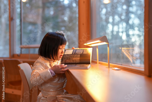 Soft light warms a quiet reading nook. Girl reads with calm focus, eyes closed. Warm glow hugs her from a small lamp. Wooden table and windows frame the scene
