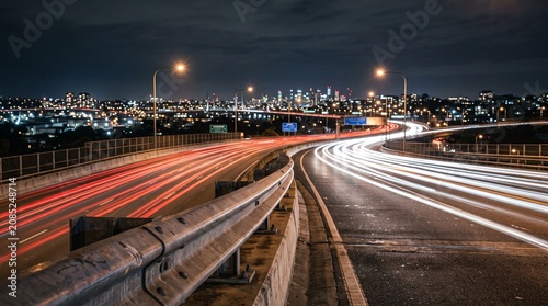 Vibrant light trails on a curved highway bridge leading to a glowing city skyline at night