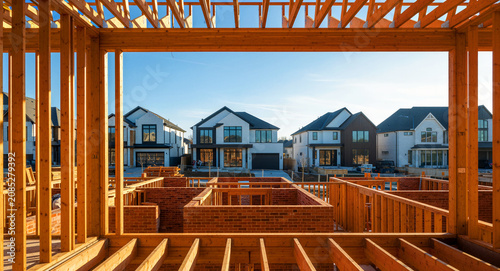 Wooden house frame view overlooking a modern suburban neighborhood development. Construction site with timber beams and luxury homes under a blue sky. Housing market and real estate growth concept.