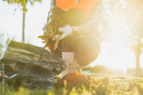 Volunteer in a safety vest collecting fallen autumn leaves into a black bag in a sunny park, participating in community cleanup and ecological conservation efforts