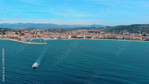 Aerial view of a passenger ferry sailing off Cannes, France, toward the old port with yachts, the Palais des Festivals, La Croisette beach, Le Suquet old town and Alpine foothills behind.