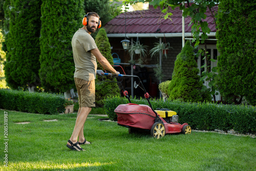 Adult man mowing green lawn with lawn mower in a well maintained backyard garden near a private house. Home gardening, lawn care, landscaping 