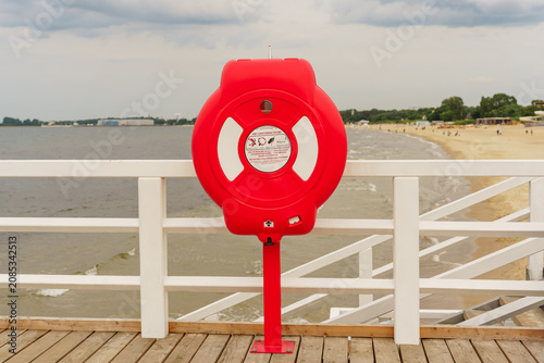Obraz na plátně Bright red lifebuoy mounted on white wooden pier railing overlooking sandy beach
