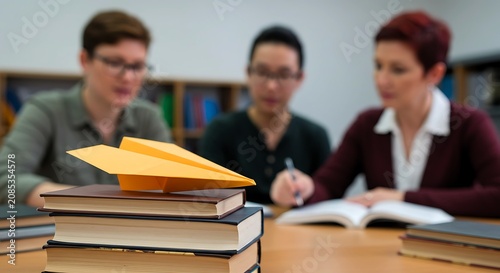 Yellow paper airplane on top of a stack of books. Diverse group of students studying in the background. Education and innovation concept in classroom. Academic learning goal development