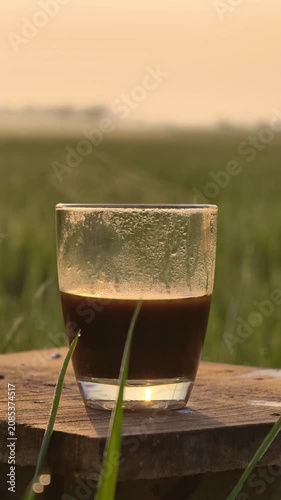 Fresh hot black coffee steaming in a glass cup on a wooden plank at sunrise with a beautiful blurred green rice field scenery background.
